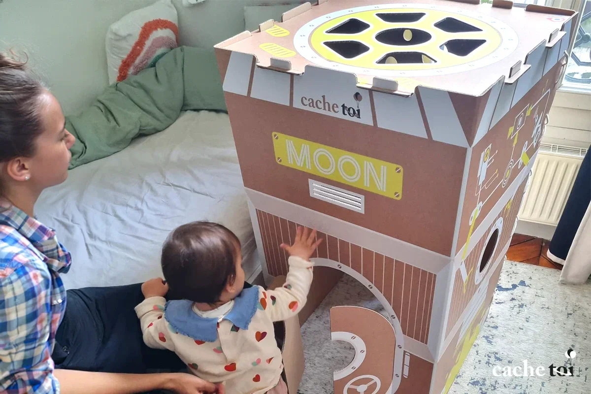 Un jeune enfant explore la cabane en carton écologique Moon de la marque cache-toi sous le regard de sa maman. Un jouet d'éveil sain, sensoriel et sans plastique pour les tout-petits.