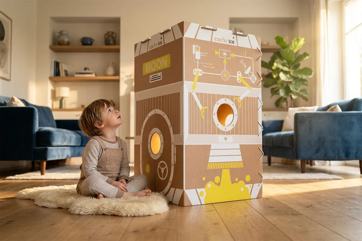 Un jeune enfant assis dans un salon lumineux à côté de la cabane Moon de cache-toi, une grande cabane en carton éco-conçue avec des découpes d'étoiles et de lune et une lumière magique à l'intérieur.