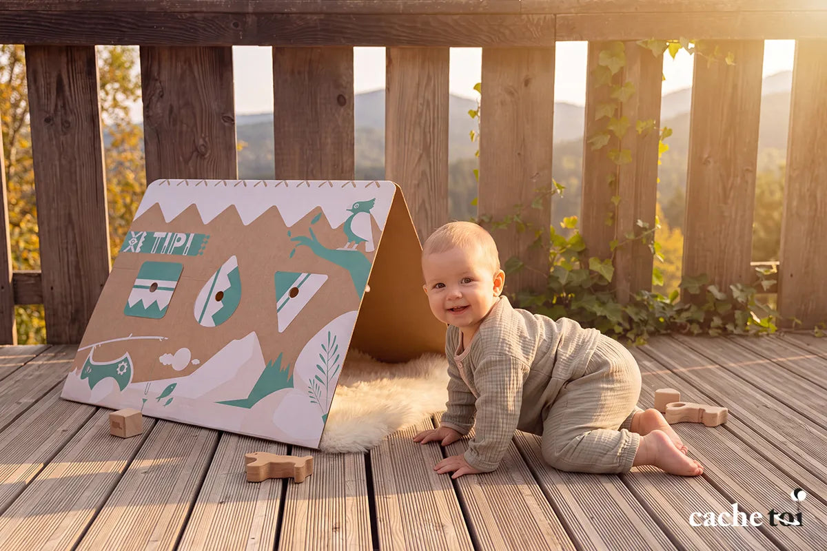 Bébé souriant à quatre pattes à côté d'une cabane en carton recyclable TIPI cache-toi posée sur une terrasse en bois ensoleillée.