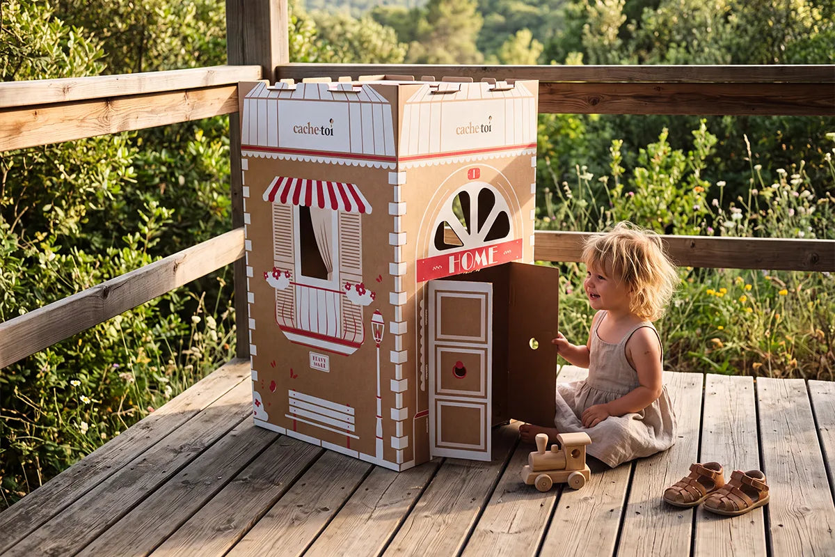 Enfant jouant avec sa cabane en carton pliable cache-toi (modèle HOME) sur une terrasse en bois. Une maisonnette zéro plastique, légère et écologique, idéale pour les vacances d'été.