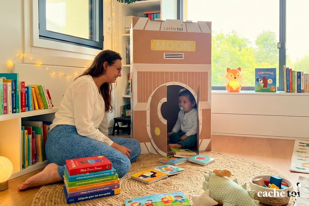 Maman partageant un moment de lecture avec son jeune enfant assis à l'intérieur de sa cabane en carton pliable modèle Fusée de la marque cache-toi.